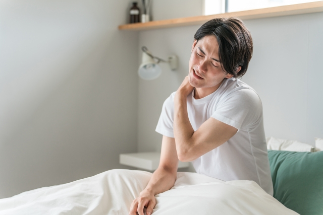 ベッドに腰かけて首の後ろを押さえる男性